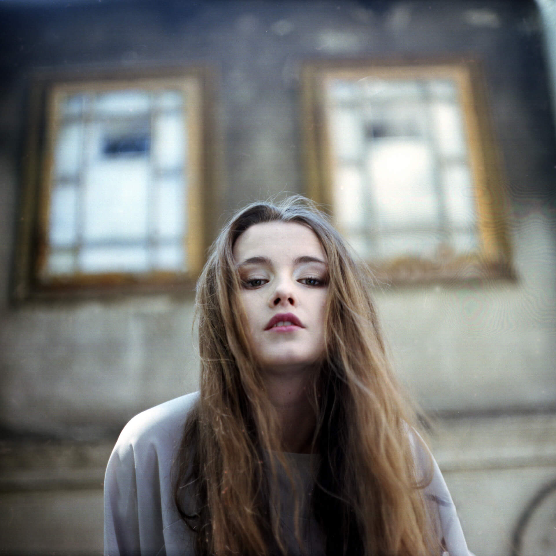 Square film portrait of the model in a pale coat in front of a shuttered facade