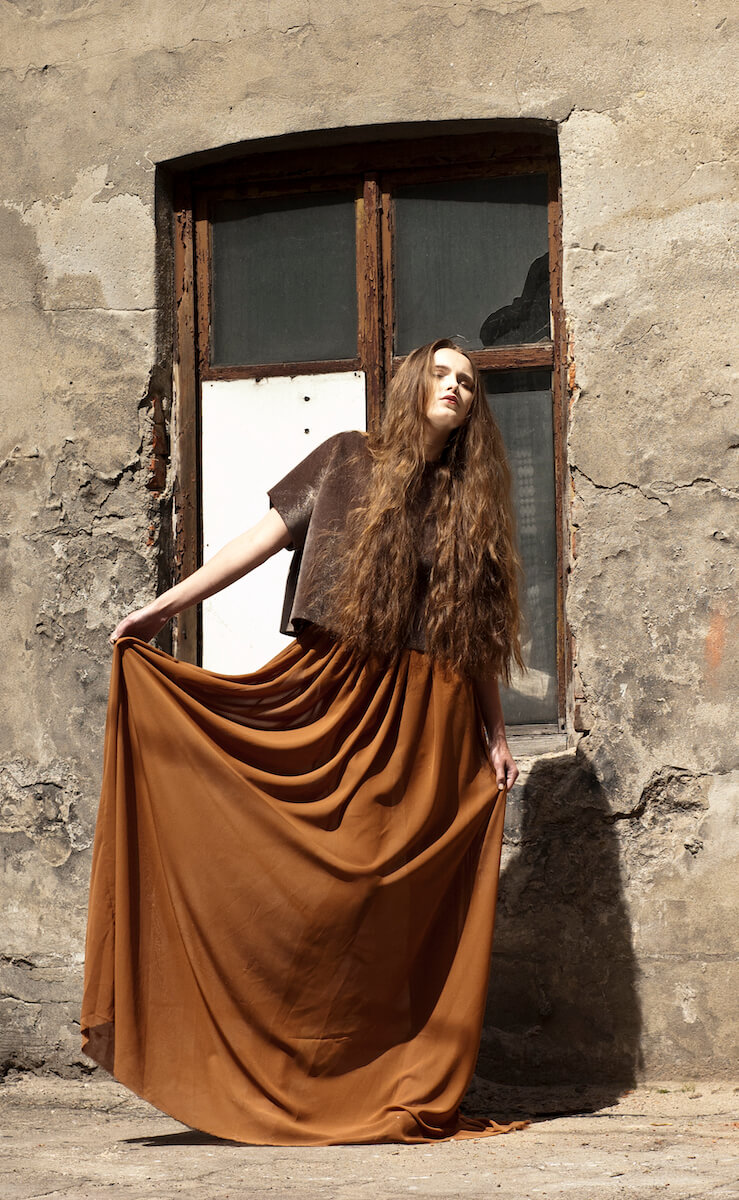 Model in a felted brown top and long rust silk skirt against a weathered wall