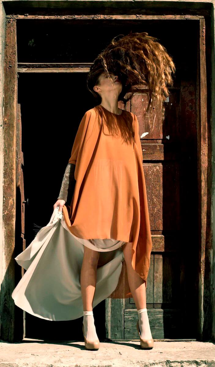 Model in an oversized rust dress over a pale silk underskirt in a doorway